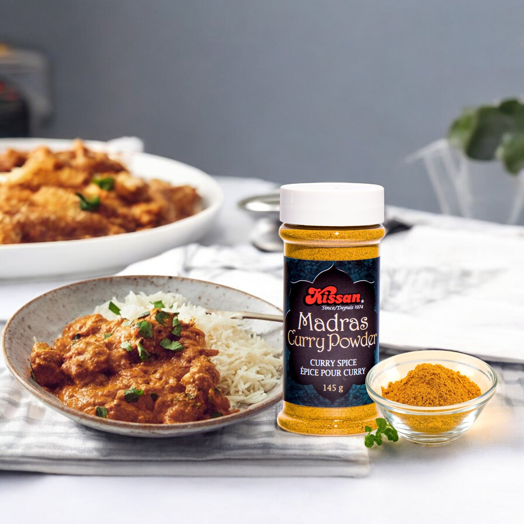 A bottle of Kissan Madras Curry Powder 145g with dish of chicken curry and rice on the side and the powder in another bowl