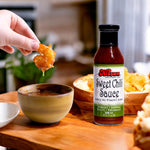 A bottle of Kissan Sweet Chili Sauce 350ml, a hand holding a shrimp dipping it in the sauce and some food in the table