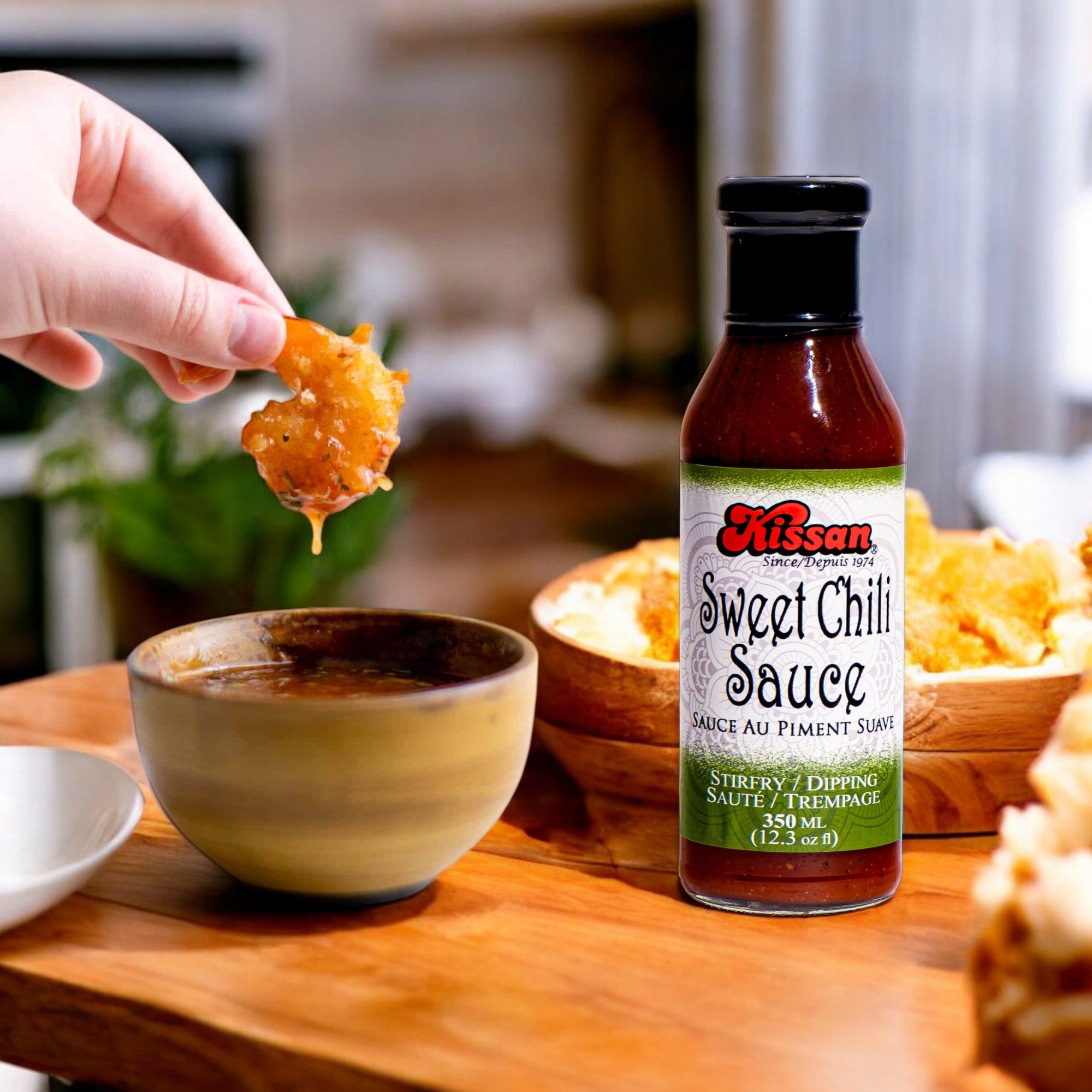 A bottle of Kissan Sweet Chili Sauce 350ml, a hand holding a shrimp dipping it in the sauce and some food in the table